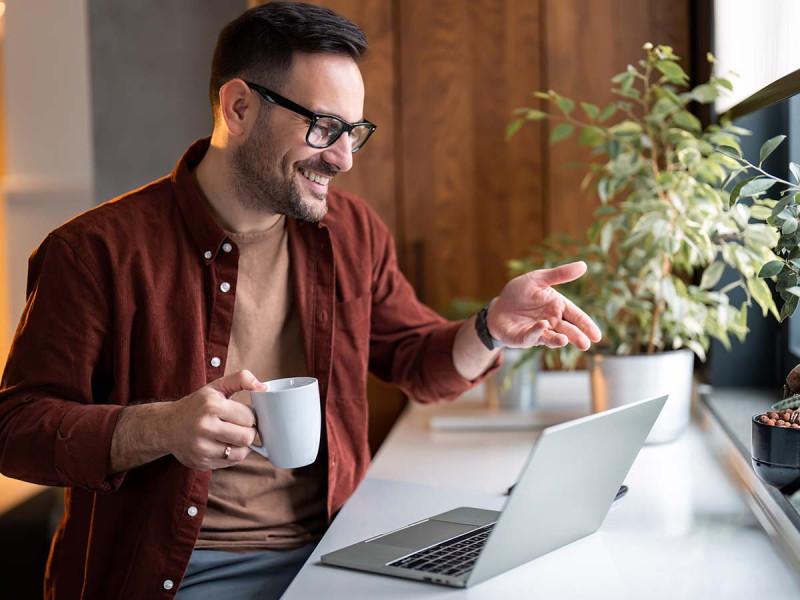 Fröhlicher, selbstbewusster Mann bei hybrider Online-Arbeit oder virtuellem Bewerbungsgespräch im Homeoffice, hält Kaffeetasse und gestikuliert während einer Videokonferenz.