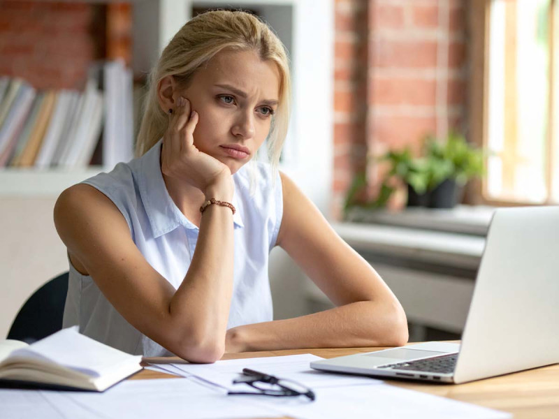 Gestresste junge Frau sitzt am Schreibtisch vor einem Laptop, wirkt erschöpft und nachdenklich, umgeben von Büchern und Arbeitsmaterialien im Homeoffice.