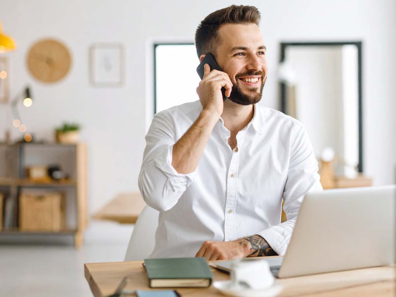 Ein junger Mann in weißem Hemd sitzt lächelnd an einem Schreibtisch, telefoniert und arbeitet an seinem Laptop. Die helle, moderne Büroeinrichtung im Hintergrund vermittelt eine freundliche Arbeitsatmosphäre.