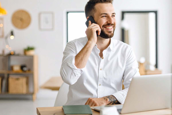 Ein junger Mann in weißem Hemd sitzt lächelnd an einem Schreibtisch, telefoniert und arbeitet an seinem Laptop. Die helle, moderne Büroeinrichtung im Hintergrund vermittelt eine freundliche Arbeitsatmosphäre.